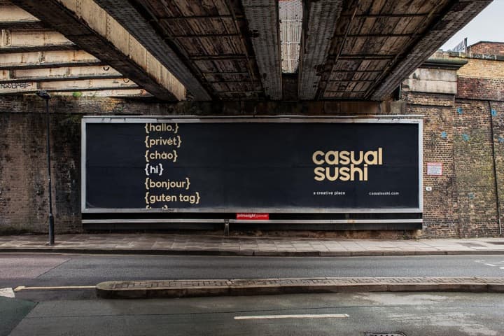A Casual Sushi billboard under a railway bridge: the agency's wordmark beside greetings in six languages